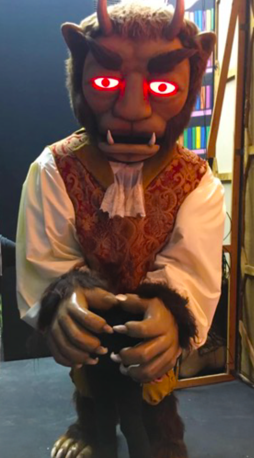 Large costume or puppet of a beast-like character with tusks, wearing a white shirt and ornate red waistcoat, posed with hands clasped together.